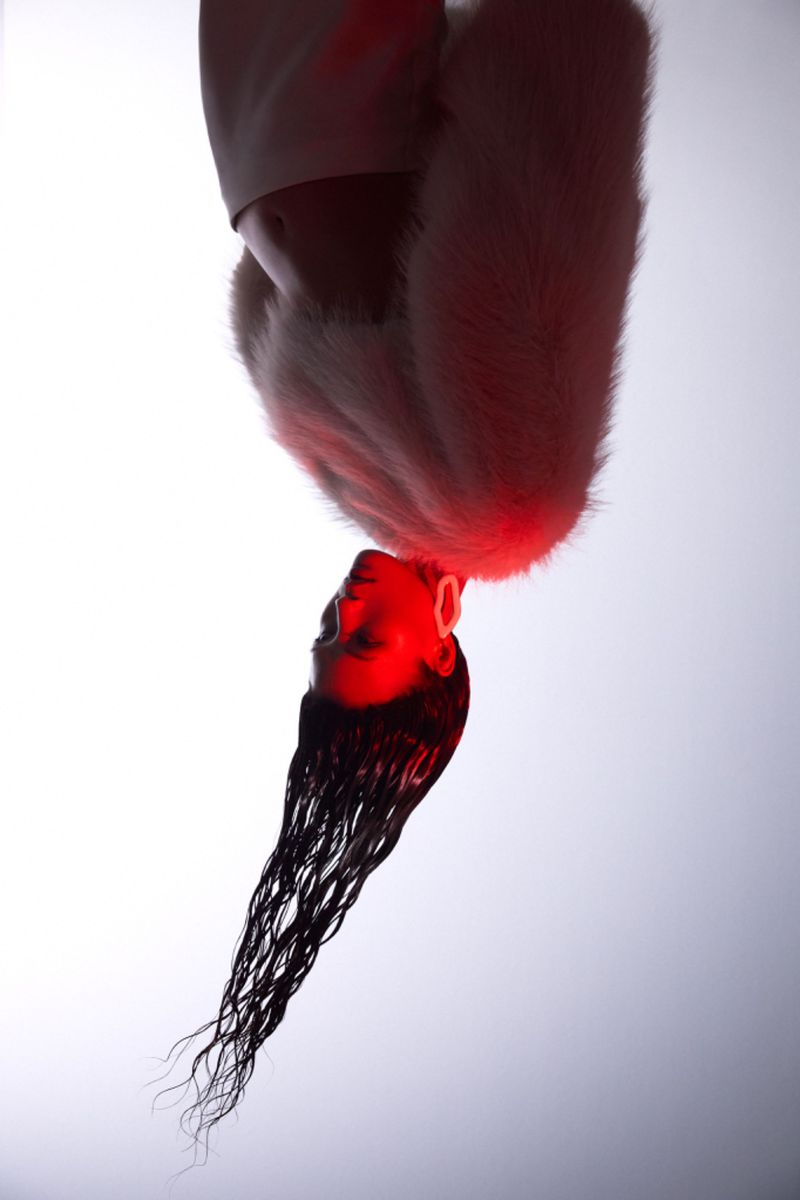 A headshot portrait of an adult woman with long dark hair, wearing a white faux fur collar, lit by soft red light against a white gradient background.