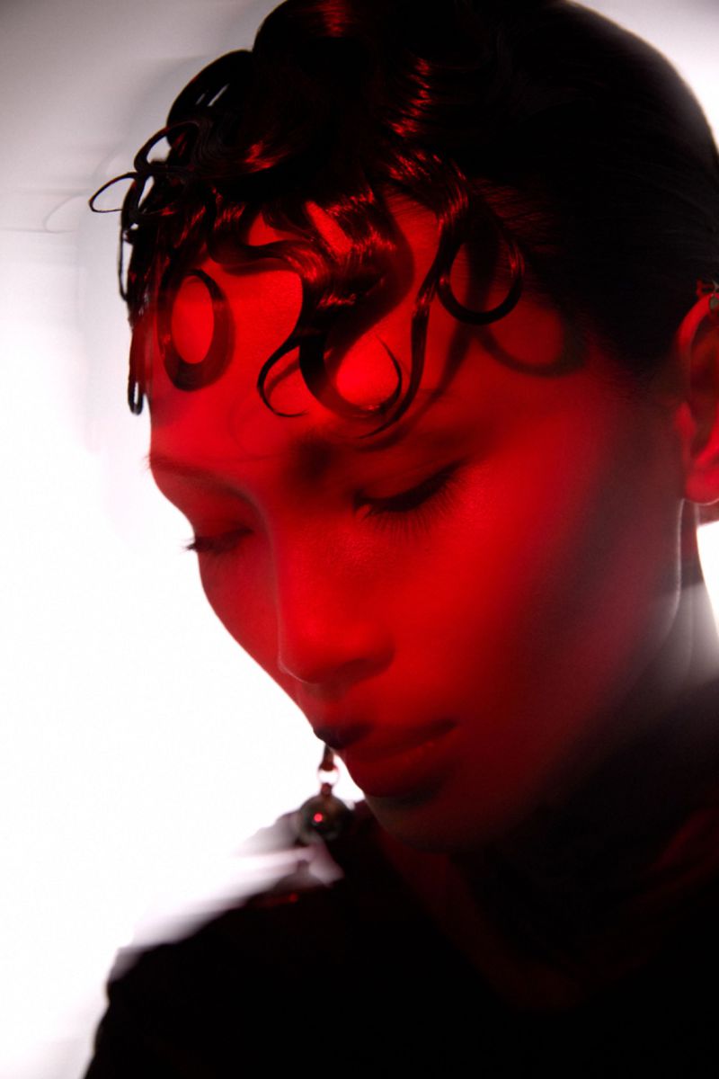 Dramatic beauty shot of a young adult woman with dark hair and an earring in front of a bright white background with red lighting.
