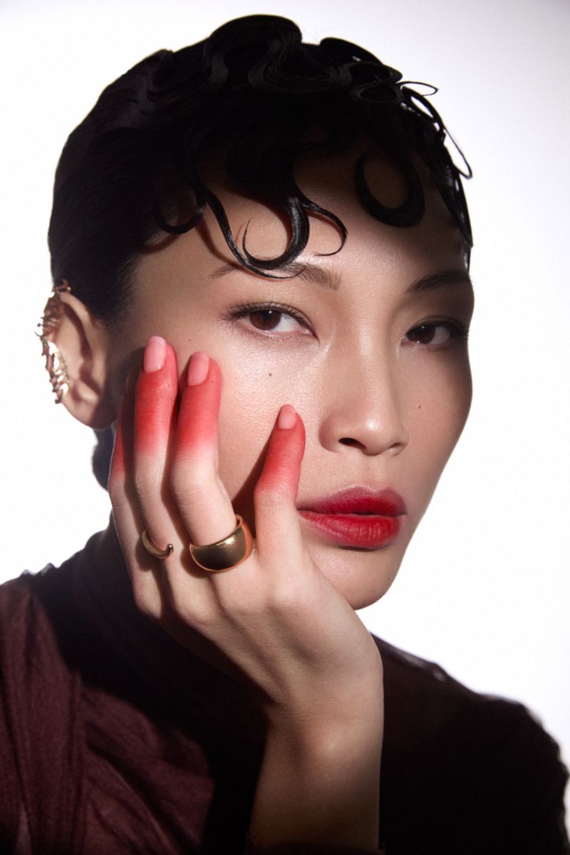 Portrait of a woman with dark curly hair, red lipstick, and red-tipped fingers against a white background in a beauty editorial style.