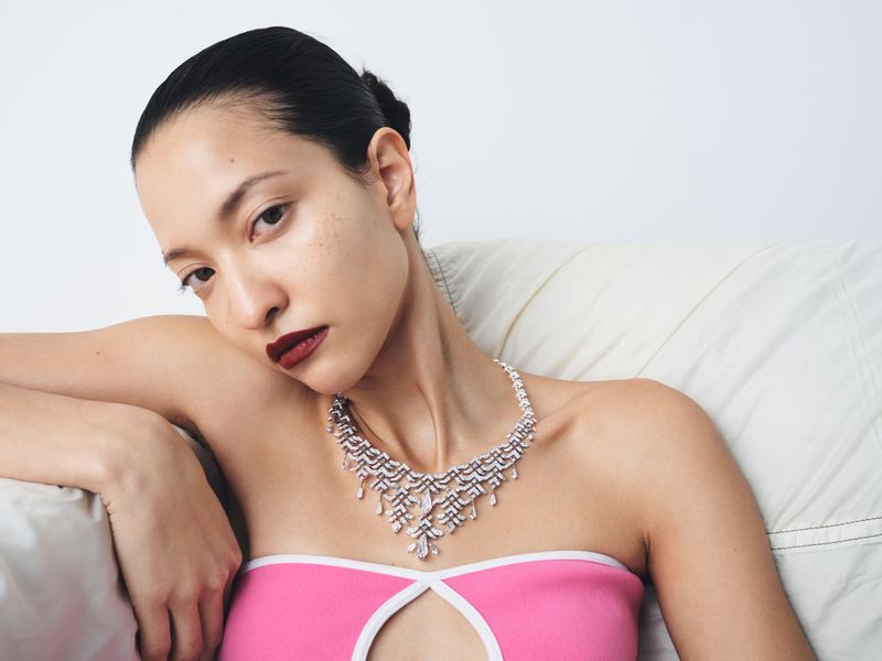 Model wearing a diamond necklace and a pink strapless top leaning against a white cushioned background