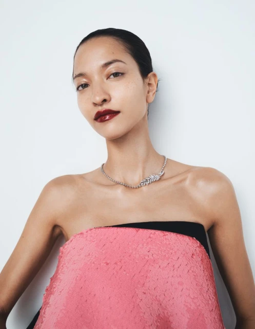 Close-up studio portrait of an adult woman with dark lipstick wearing a pink sequin dress and a silver necklace against a white background.