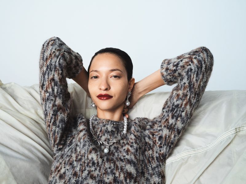 Elegant woman with dark hair poses in a mottled brown sweater on a light couch, wearing pearl jewelry and dark red lipstick in an indoor setting.