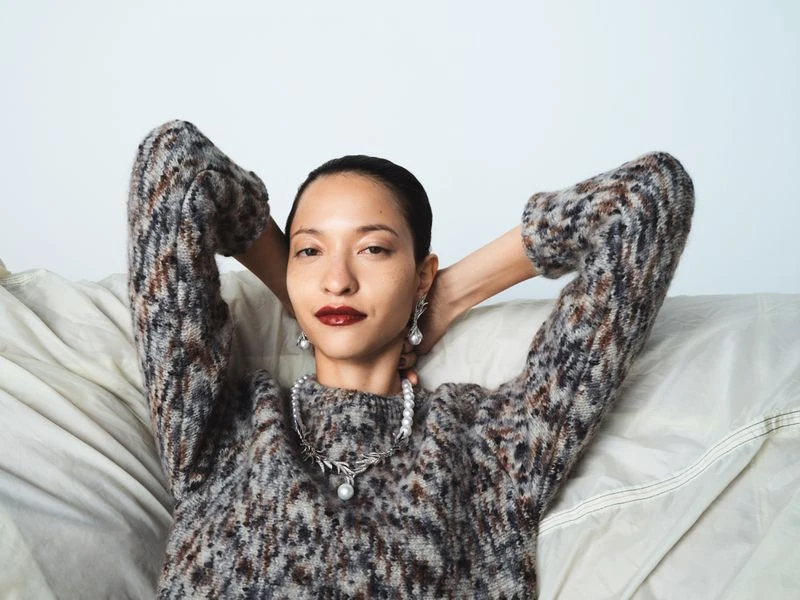 Elegant woman with dark hair poses in a mottled brown sweater on a light couch, wearing pearl jewelry and dark red lipstick in an indoor setting.