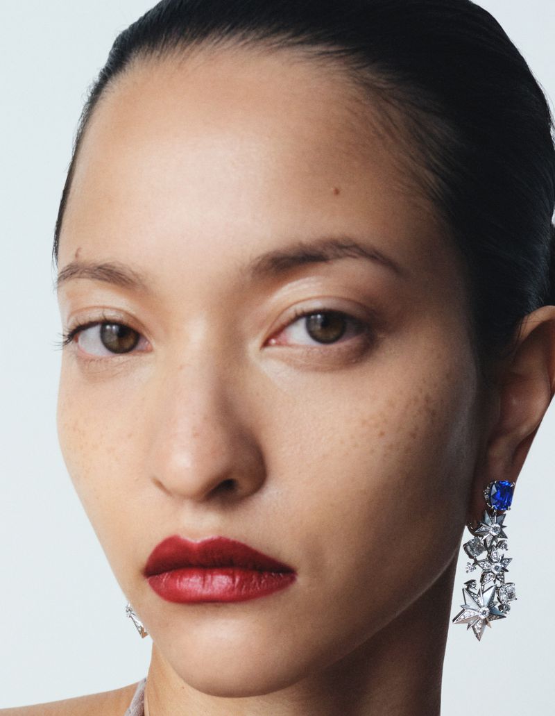 Close-up shot of a young adult woman with brown hair pulled back, wearing dark red lipstick and dangling blue and silver earrings.