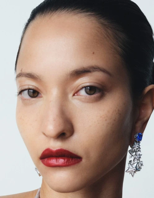 Close-up shot of a young adult woman with brown hair pulled back, wearing dark red lipstick and dangling blue and silver earrings.