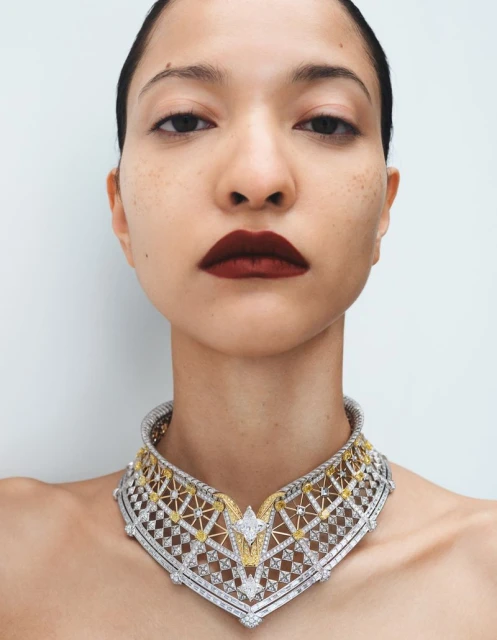 Close-up studio shot of a woman with fair skin and dark lipstick, wearing an ornate, diamond-studded necklace against a plain white background.