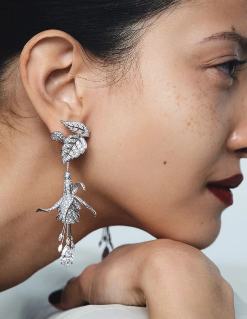Close-up of an adult female wearing a pair of white gemstone flower and leaf drop earrings with dark red lipstick against a white backdrop.