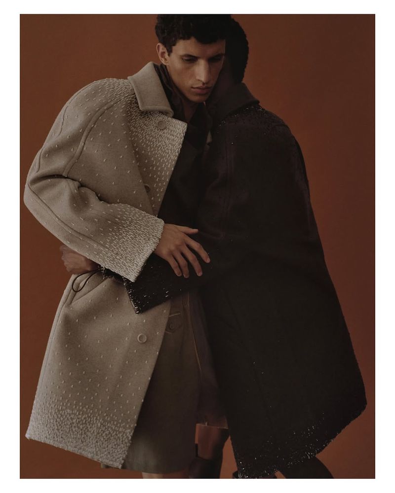 Two men in a close embrace, showcasing contrasting overcoats in a studio setting with a warm brown background, highlighting texture and form.