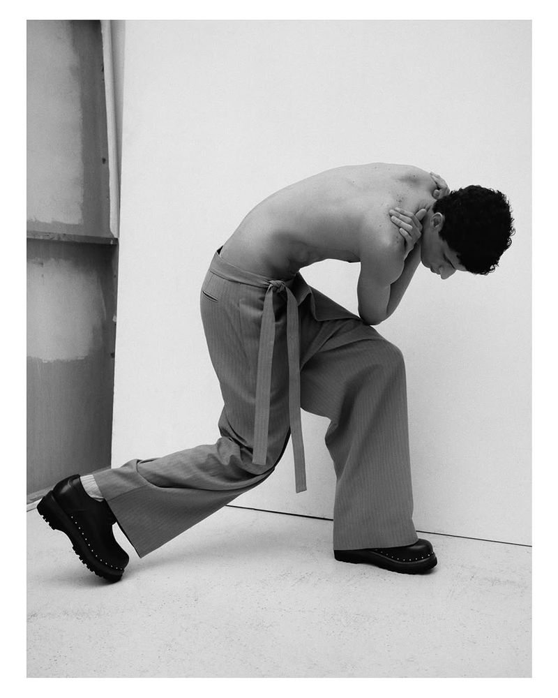 A bare-chested man in loose pants poses in a studio with an arched back and crossed arms, standing next to a textured wall.