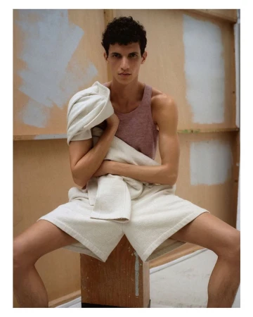 Man poses in a photo studio wearing a tank top and shorts, draped with a towel, sitting on a wooden stool against a neutral backdrop.