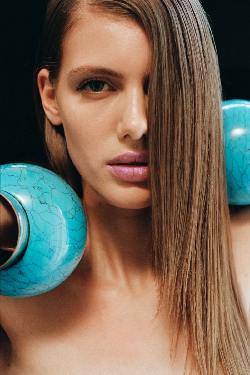 Close-up portrait of a light-skinned woman with long, light brown hair, partially obscuring her face, with blue-green vases.