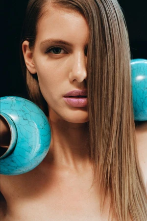 Close-up portrait of a light-skinned woman with long, light brown hair, partially obscuring her face, with blue-green vases.
