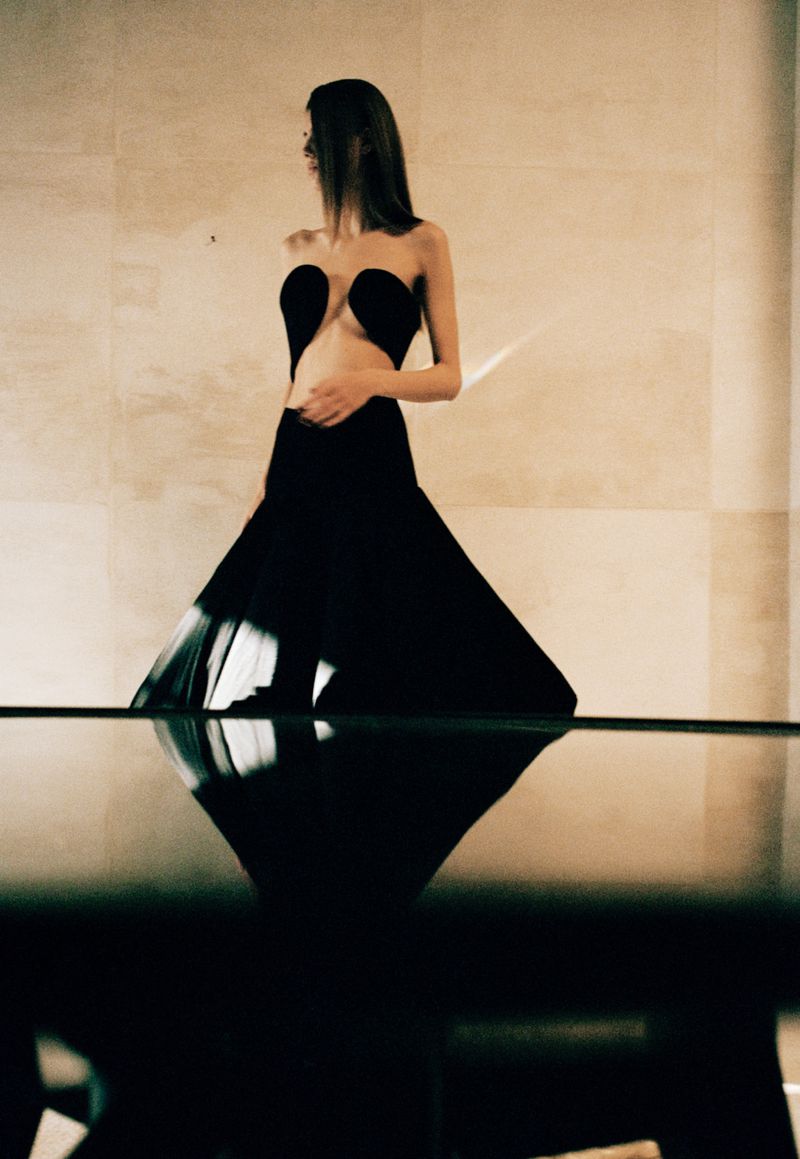 Elegant woman in a black, strapless gown with a cut-out bodice, looking over her shoulder in a minimalist indoor setting. Fashion photoshoot.