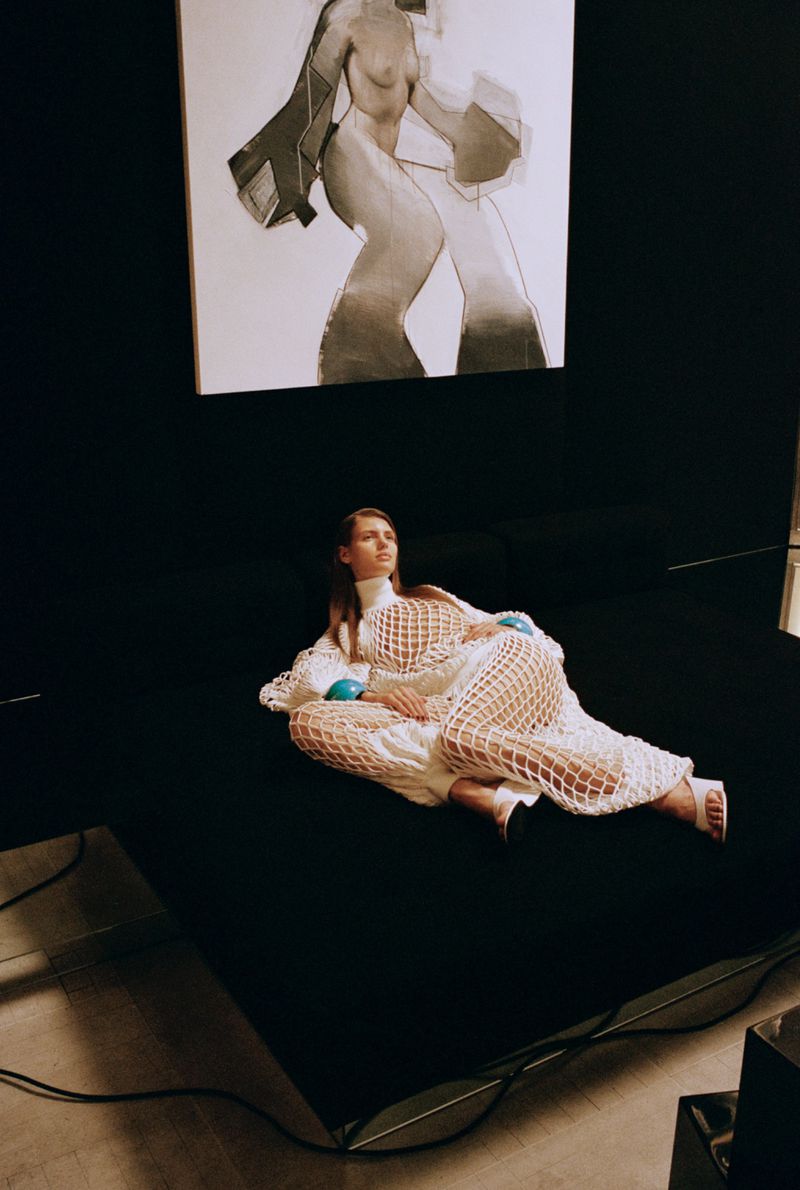 Fashion shot of a woman in a white crocheted dress reclining on a black surface, with a monochromatic artwork in the background.