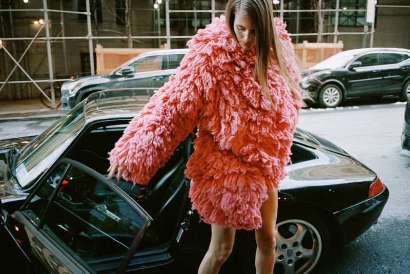 Woman in a pink coat getting into a black sports car on a city street with buildings and traffic in the background on an overcast day.