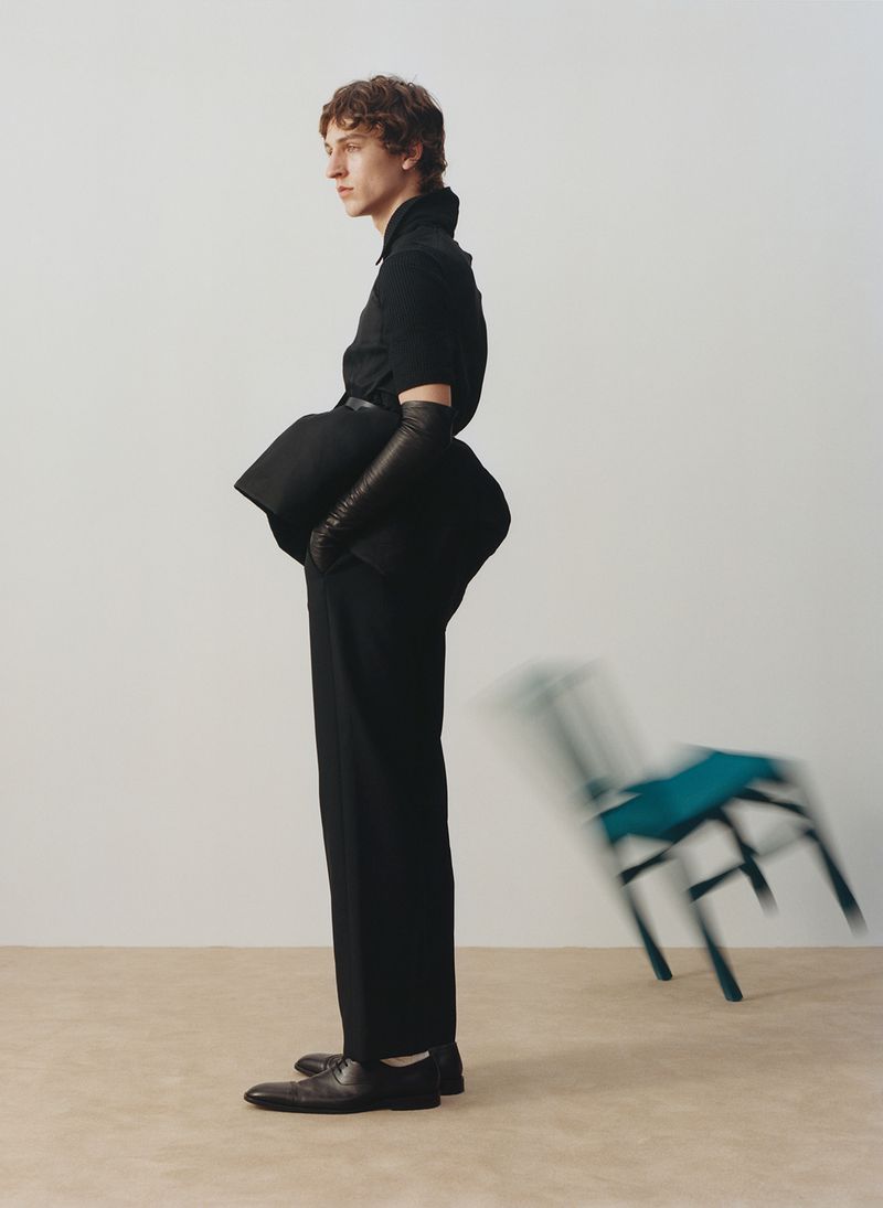 A slender man in a black suit and leather gloves strikes a side pose in a studio setting, with a blurred teal chair in the background.