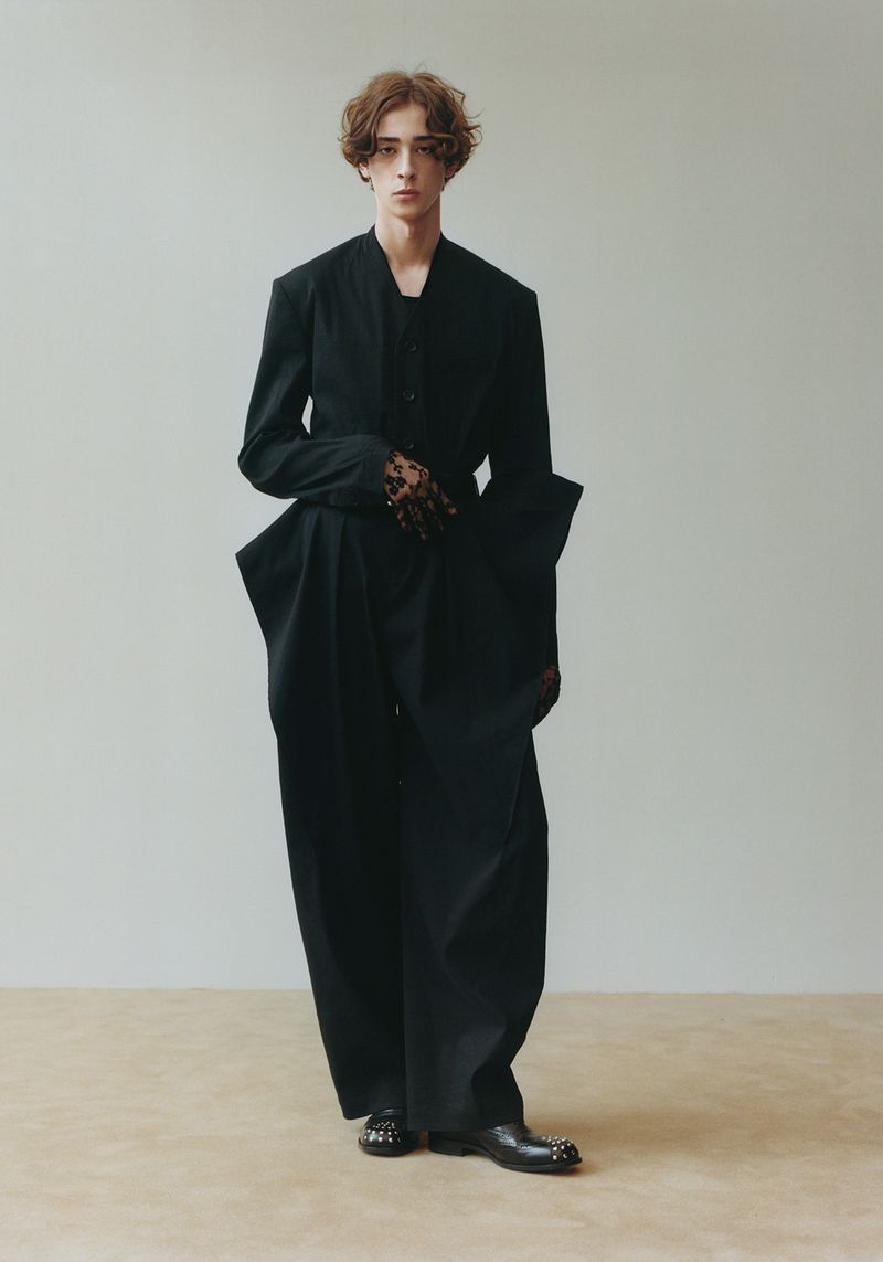 Full-length portrait of a young adult male fashion model in a black suit with wide-legged pants standing against a neutral background.
