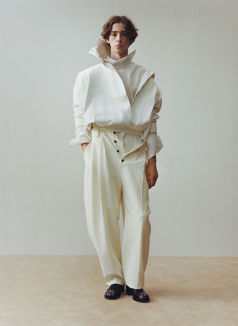 Full-length portrait of a male fashion model with medium brown hair in a white, asymmetrical shirt and pants against a neutral backdrop.