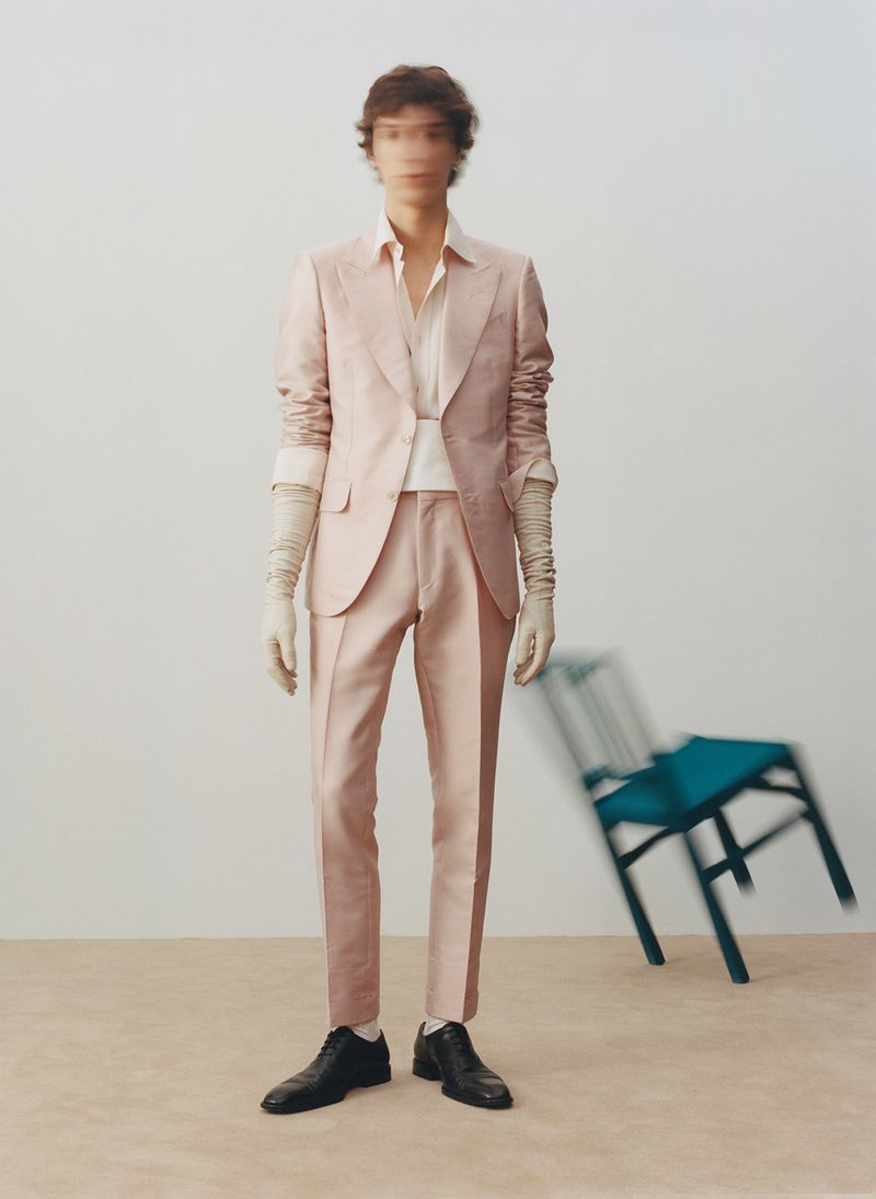 A blurry male model in a blush suit stands indoors with a teal chair, shot in a medium full shot in a minimalist fashion editorial.