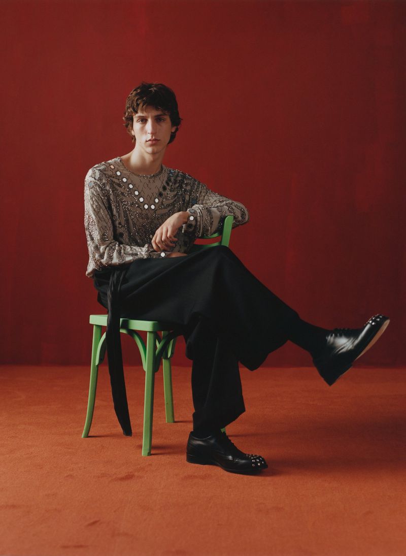 Adult male seated on a green chair in a photo studio wearing a patterned top, black culottes and studded black shoes. Red backdrop.