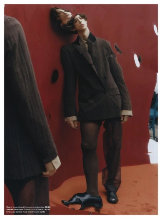 Model in brown blazer and sheer trousers posing against a vibrant red backdrop for AnOther Man Magazine.