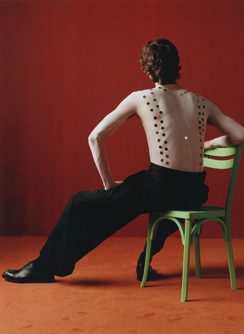 Model seated backwards on a green chair with painted white back and circular dots, wearing black trousers and shoes against a red background.