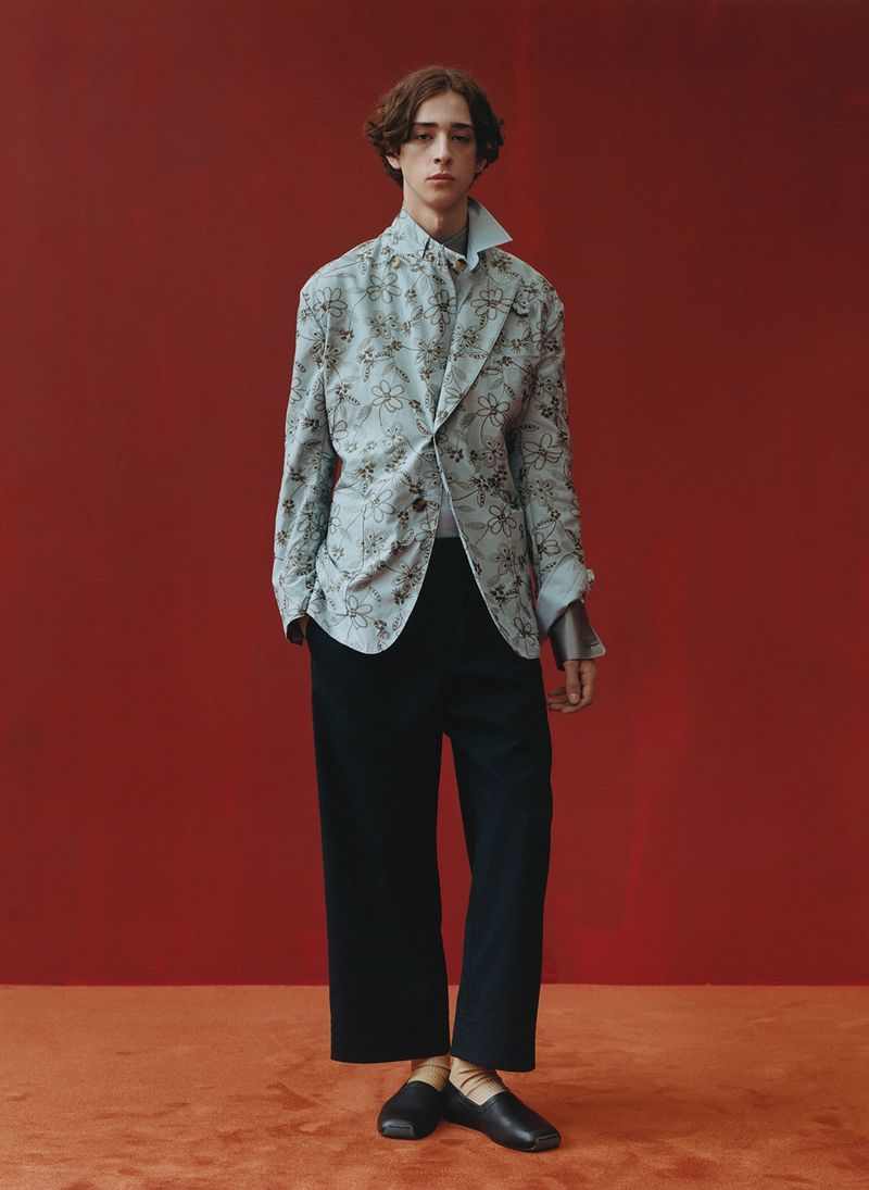 Full-length shot of a young man in a patterned blazer, collared shirt, black pants, and leather shoes against a red backdrop.