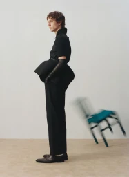 Model standing in profile wearing black attire with long leather gloves against a plain backdrop with a blurred moving chair