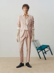 Model in pink suit and gloves with motion blur face standing before a pale wall next to a falling chair
