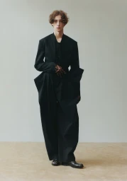 A male model wearing a tailored black suit stands against a plain neutral background in an editorial photograph.
