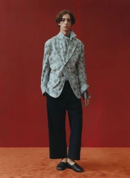 A male model wearing a patterned grey blazer and dark trousers standing against a textured dark red wall.