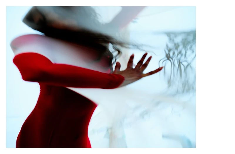 Woman in a red bodysuit poses behind a translucent white sheet, hand extended toward the viewer in a studio environment.