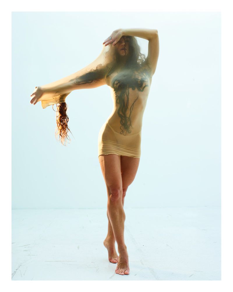 A woman in a short dress stands in a photo studio with sheer fabric draped over her, holding one arm up and the other extended.