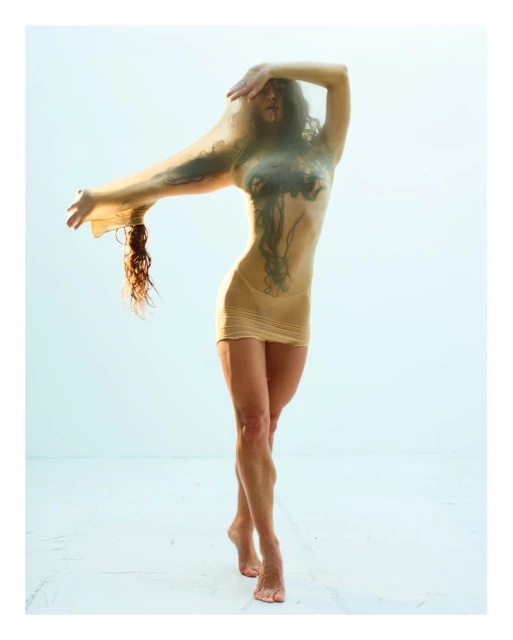 A woman in a short dress stands in a photo studio with sheer fabric draped over her, holding one arm up and the other extended.