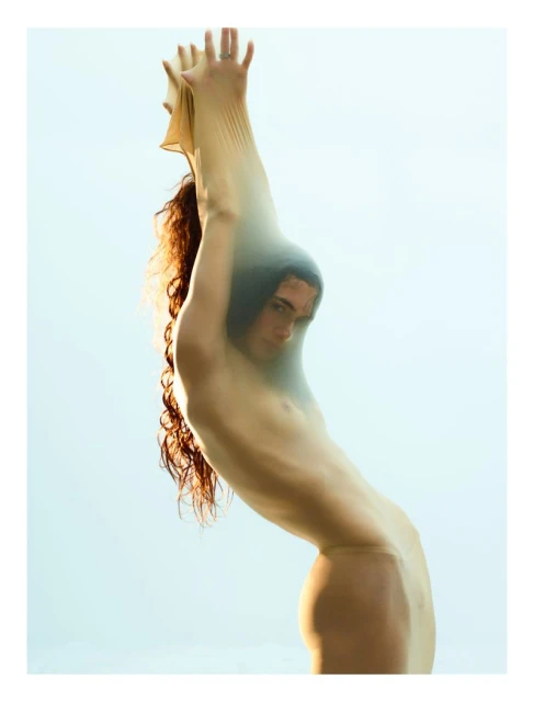Artistic nude portrait of a woman with long curly hair in a sheer fabric against a light blue background in a photo studio.