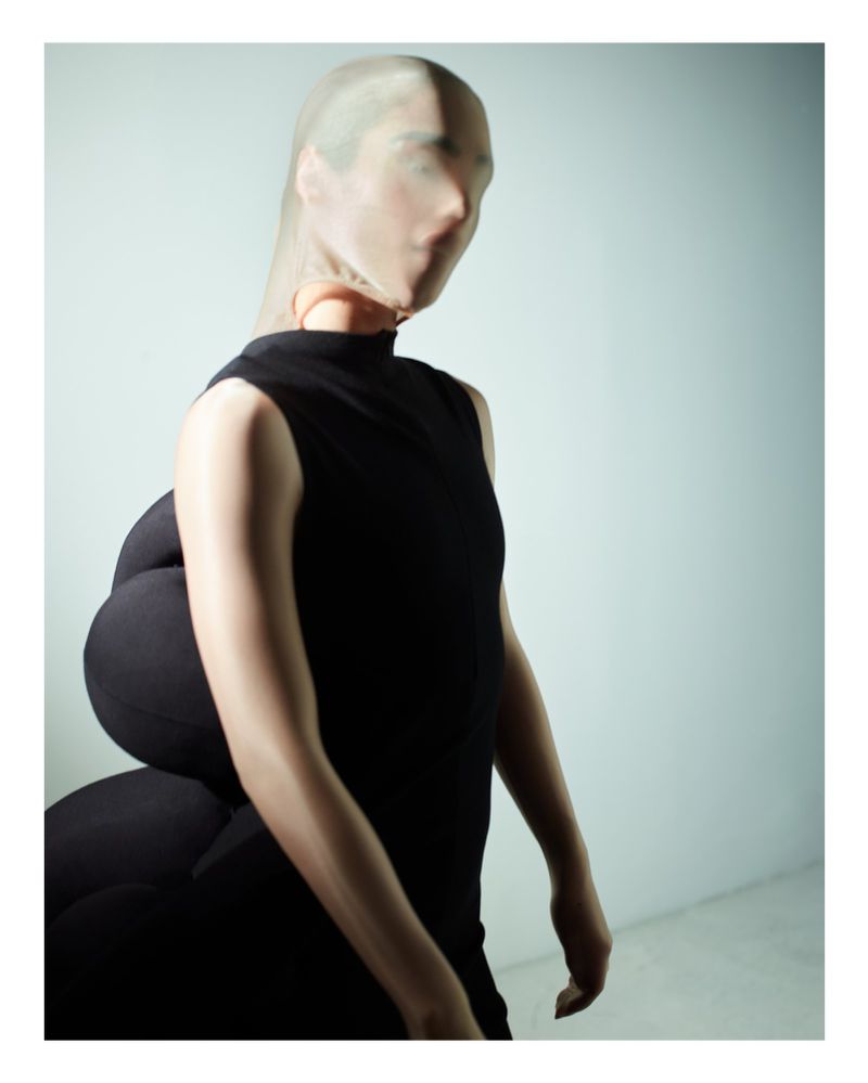 Woman in black dress and sheer stocking mask poses in studio with soft lighting creating surreal fashion editorial with unique silhouette.