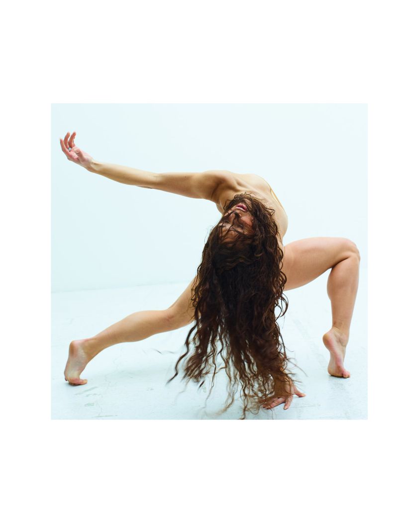 Female dancer in studio, strikes expressive pose, back arched, arms reaching, long dark hair flowing, wearing minimal clothing against white background.