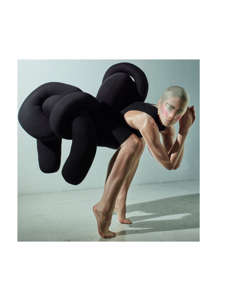 Person in creative fashion pose with black knot-like design, wearing a nude bodysuit, hands on head, and neutral facial expression.
