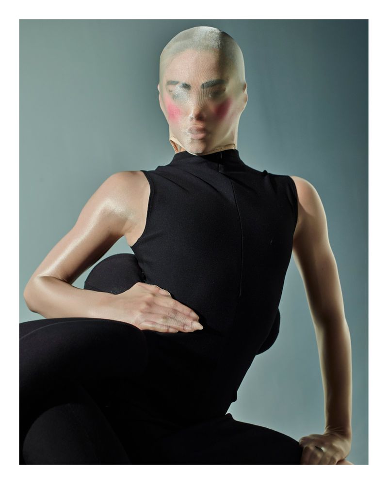 Studio shot of a woman in a black sleeveless top and pants, posing with a nylon stocking over her head against a blue background.