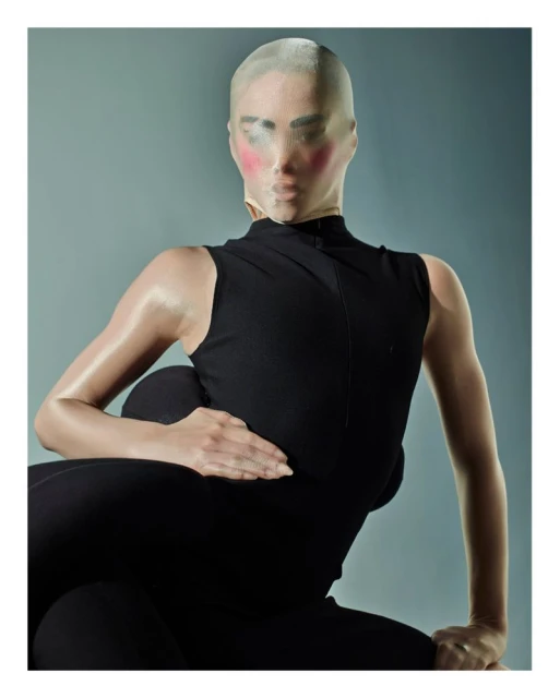 Studio shot of a woman in a black sleeveless top and pants, posing with a nylon stocking over her head against a blue background.