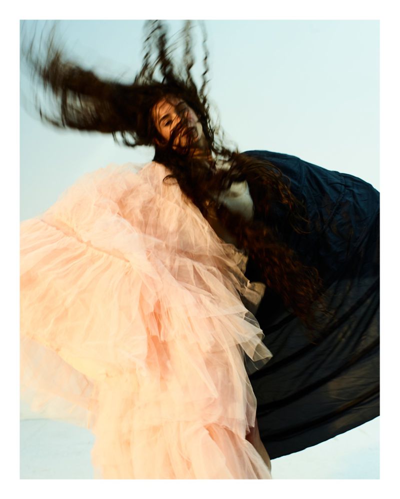 A person with long brown hair and a pink tulle dress and a black top is in motion against a light blue background in a studio shot.