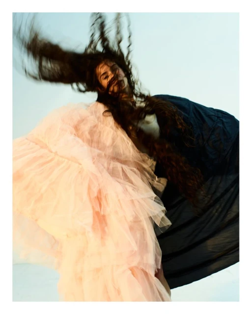 A person with long brown hair and a pink tulle dress and a black top is in motion against a light blue background in a studio shot.