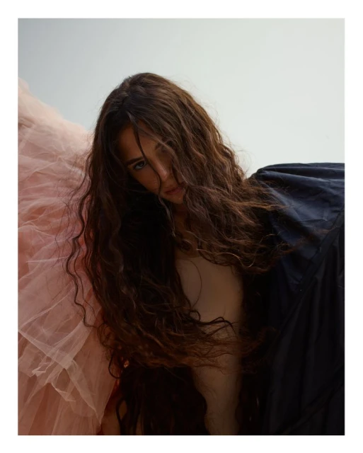 Portrait of a woman with long dark hair wearing a dark blue top, with a soft pink tulle-like fabric in the background.