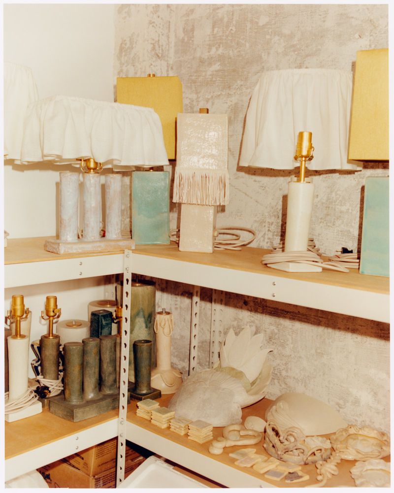 Assorted lamps and lamp parts on metal shelves in a storage room, featuring varying designs and materials, under soft, neutral lighting.
