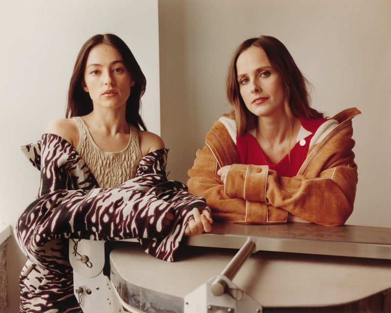 Two young women pose indoors wearing contemporary fashion. One wears a patterned coat and the other a tan jacket and red top in a studio setting.