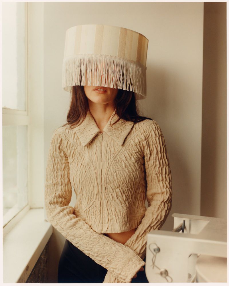 Woman wears lampshade hat in a fashion photograph. She wears a beige textured top, stands next to a window and sewing machine.