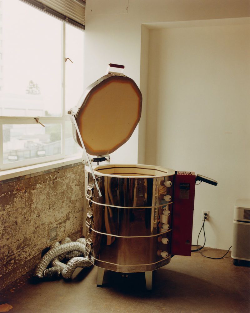An electric pottery kiln is set up indoors near a window, with silver vent tubing running along the floor in a studio setting.