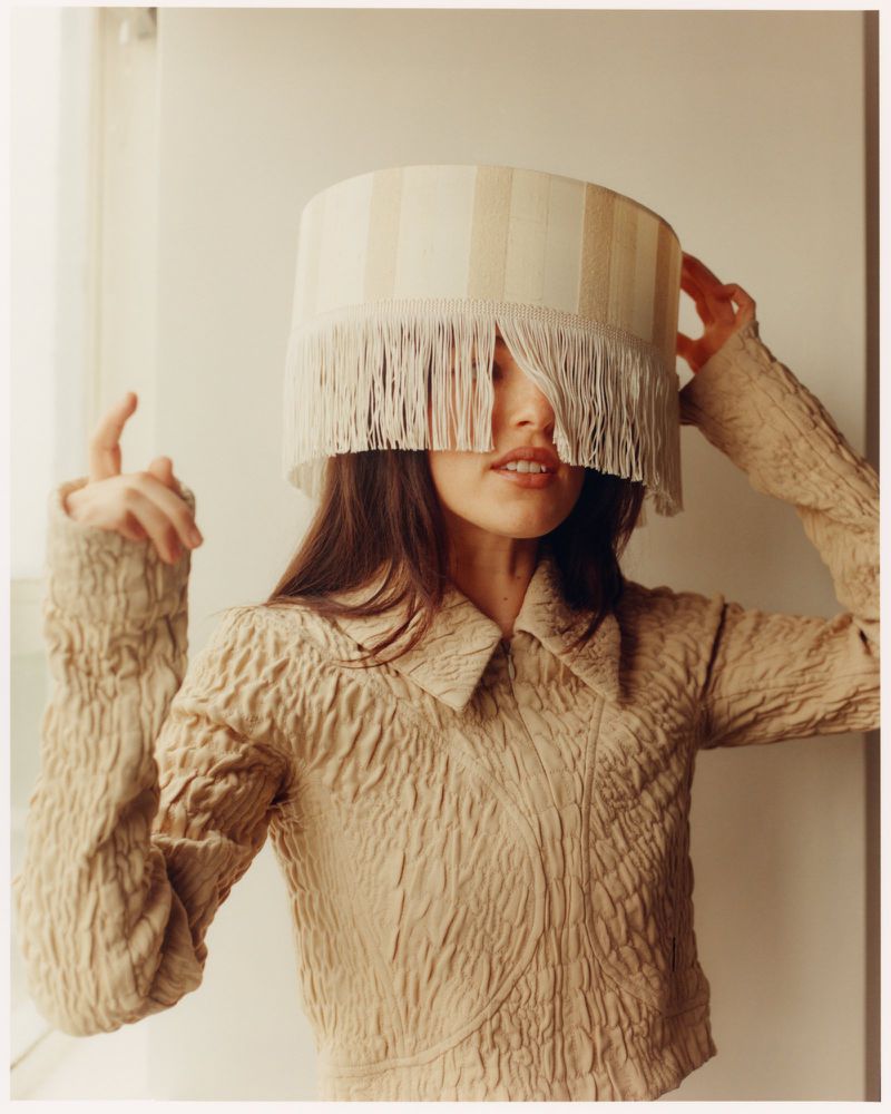 Woman poses indoors wearing fringed lampshade as hat and collared crop-top, with light and shadow from window nearby.
