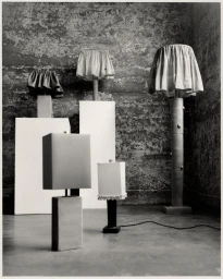 Monochrome still life of several handcrafted sculptural lamps and pedestals against a textured concrete wall.