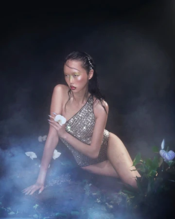A woman in a sparkly swimsuit poses in a misty, dreamlike environment surrounded by greenery, holding a white orchid flower.